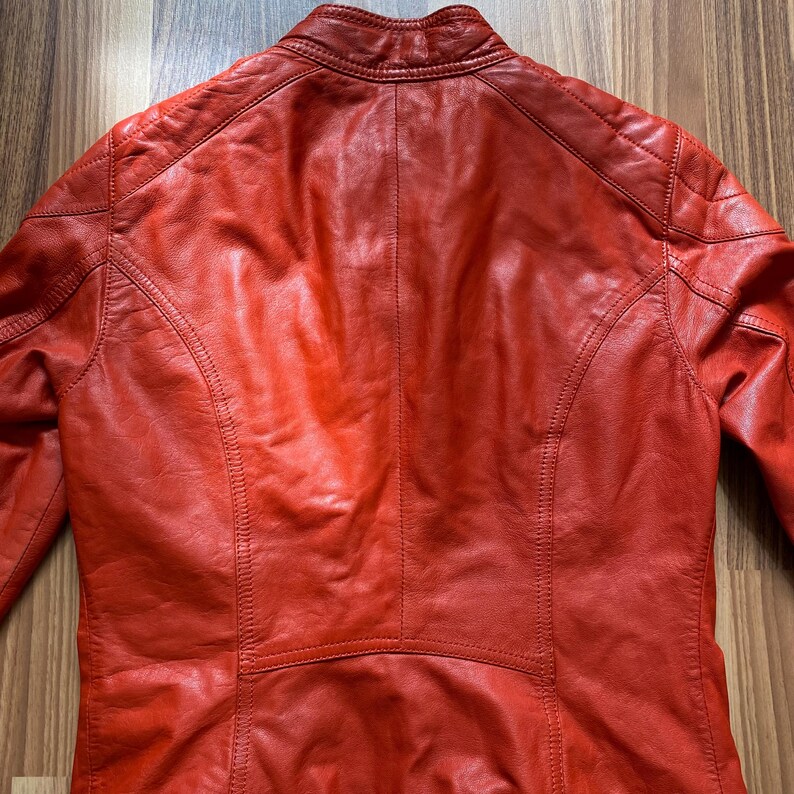 May include: A vibrant red leather jacket, viewed from the back. The jacket features a stand-up collar, quilted shoulder detailing, and a tailored fit. The leather has a slightly distressed appearance, with visible stitching and panel seams.