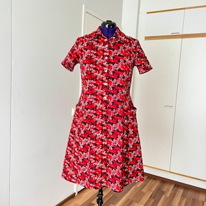 May include: A knee-length dress in a vibrant red fabric featuring a dense, repeating pattern of small black and white floral shapes. The dress has short sleeves, a classic collar, a full-length front button closure, and two large patch pockets.