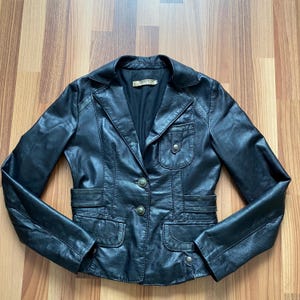 K&ouml;nnte beinhalten: Schwarzer Lederblazer mit Reverskragen, zwei Vordertaschen und zwei Brusttaschen. Der Blazer hat einen Zwei-Knopf-Verschluss und dekorative Kn&ouml;pfe an den Taschen. Die Jacke wird auf einer Holzoberfl&auml;che pr&auml;sentiert.