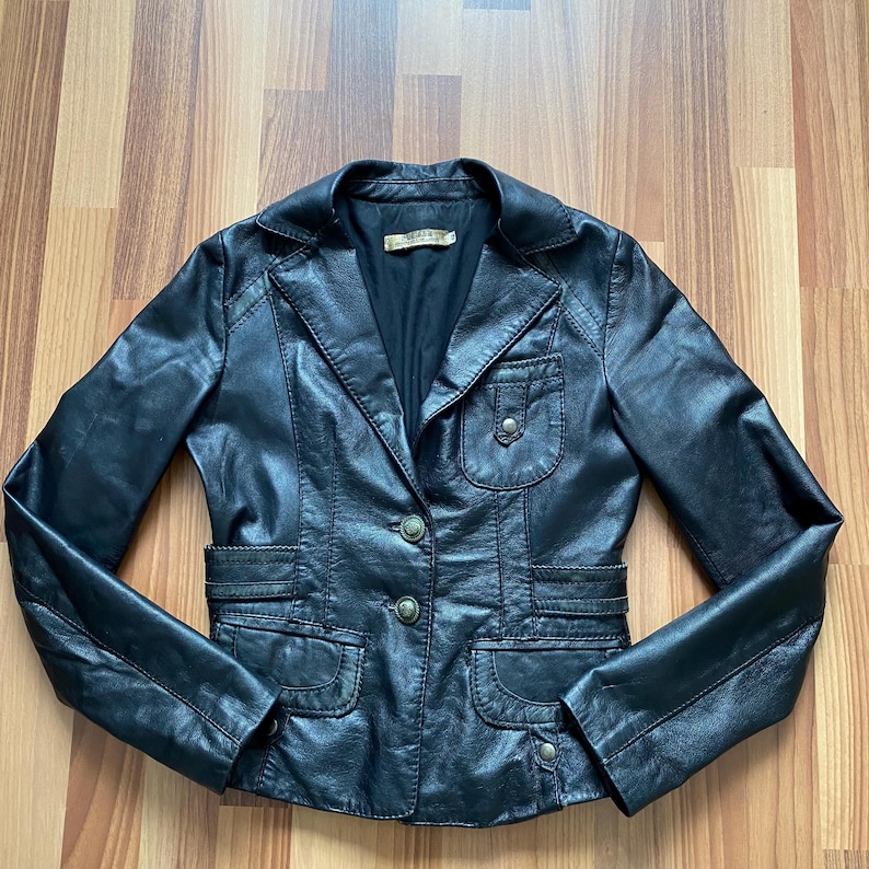 K&ouml;nnte beinhalten: Schwarzer Lederblazer mit Reverskragen, zwei Vordertaschen und zwei Brusttaschen. Der Blazer hat einen Zwei-Knopf-Verschluss und dekorative Kn&ouml;pfe an den Taschen. Die Jacke wird auf einer Holzoberfl&auml;che pr&auml;sentiert.
