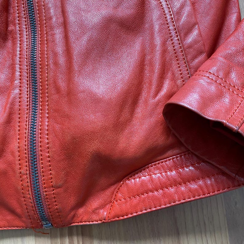 May include: Close-up of a red leather jacket with a silver zipper. The jacket has detailed stitching along the seams and around the cuffs. The leather has a slightly textured appearance.