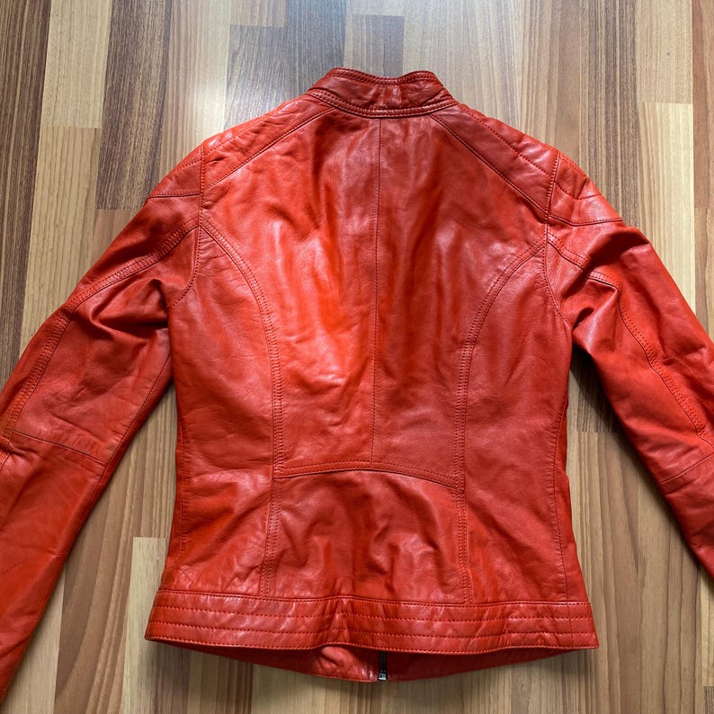 May include: A red leather jacket with a stand-up collar and quilted shoulder detailing. The jacket has a fitted silhouette and visible stitching. The back of the jacket is visible, showcasing its design and construction. The jacket is made of leather.