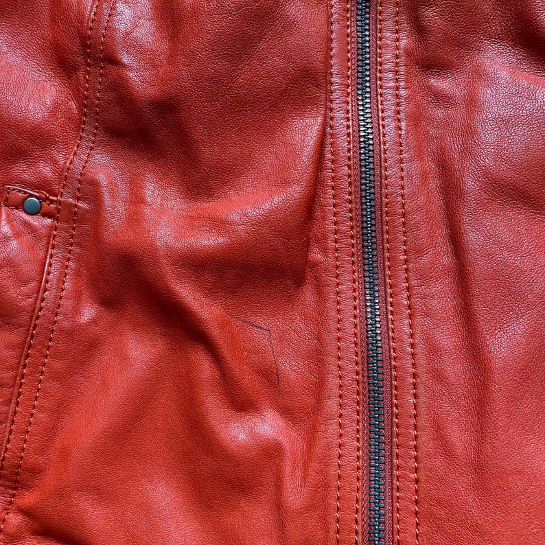 May include: Close-up of a red leather jacket with a black zipper. The jacket has visible stitching and a small pocket detail with a silver button. The leather has a textured appearance.