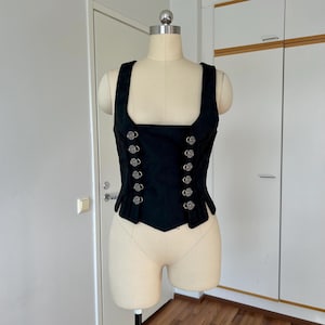 May include: A black vest with a square neckline and silver buttons. The vest is displayed on a mannequin. The buttons are arranged in two vertical rows. The vest has a fitted shape and a slightly pointed hemline.