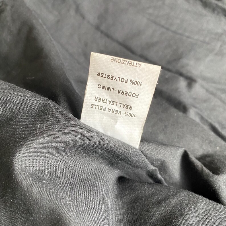K&ouml;nnte beinhalten: Nahaufnahme des Inneren eines schwarzen Kleidungsst&uuml;cks mit einem wei&szlig;en Etikett. Das Etikett gibt an, dass das Futter aus 100 % Polyester und das Echtleder aus 100 % Vera Pelle besteht. Das Wort "ATTENZIONE" ist ebenfalls sichtbar.