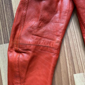 May include: Close-up of a section of a bright red leather jacket. The leather has a textured appearance with visible stitching along the seams. The jacket appears to be well-worn, with some creases and subtle variations in color.