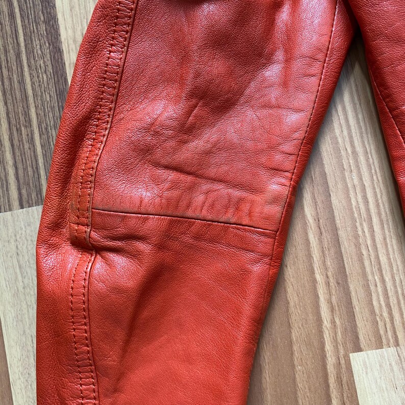 May include: Close-up of a section of a bright red leather jacket. The leather has a textured appearance with visible stitching along the seams. The jacket appears to be well-worn, with some creases and subtle variations in color.
