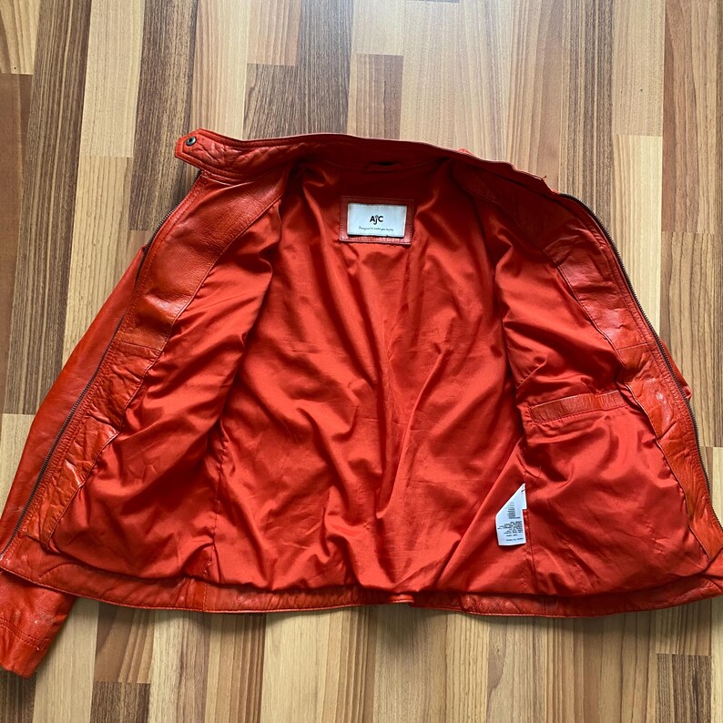 May include: Open orange leather jacket with a matching orange satin lining. The jacket has a classic design with a collar, zipper, and pockets. The label inside the jacket reads "AJC".