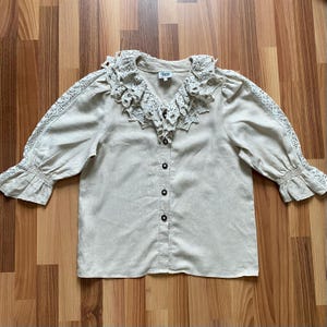 May include: A beige button-down blouse featuring a large, ruffled crochet collar and matching crochet lace panels running down the length of the sleeves. The garment has three-quarter length sleeves with gathered, elasticized cuffs and dark buttons.