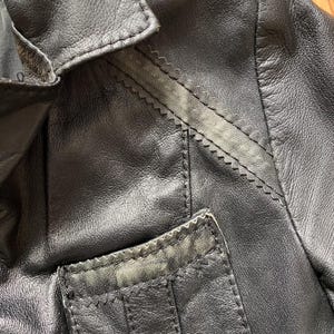 K&ouml;nnte beinhalten: Nahaufnahme einer schwarzen Lederjacke. Die Jacke hat einen Reverskragen, eine kleine Tasche und dekorative N&auml;hte entlang der N&auml;hte. Das Leder scheint abgenutzt zu sein, wobei einige Bereiche eine hellere Farbe aufweisen.