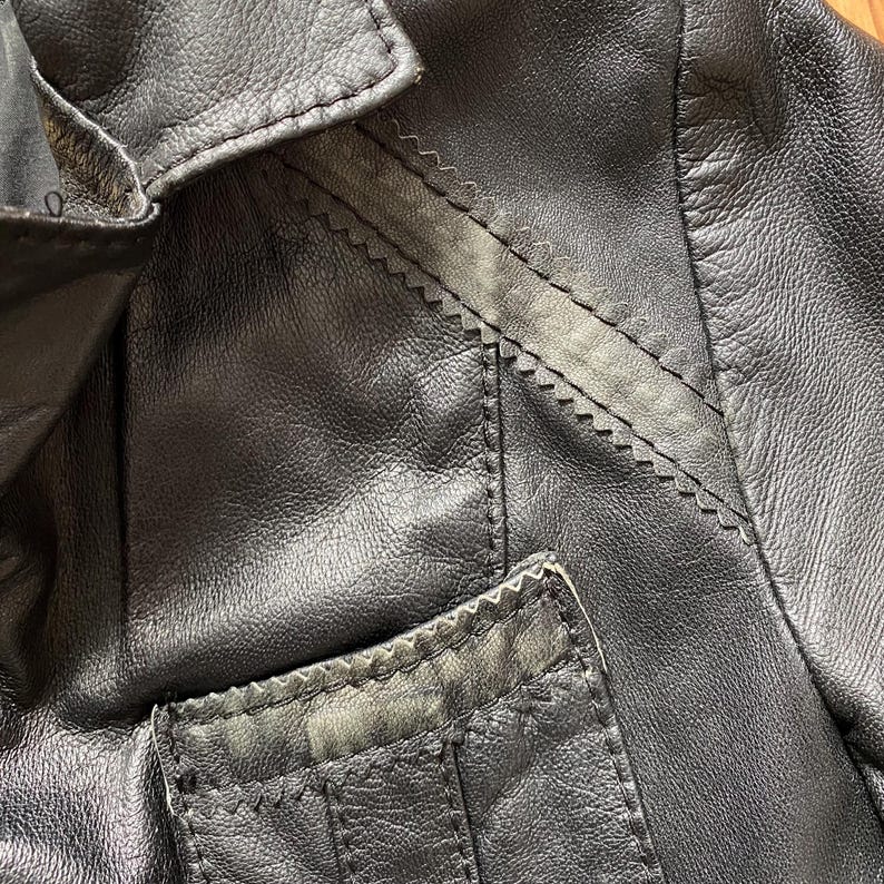 K&ouml;nnte beinhalten: Nahaufnahme einer schwarzen Lederjacke. Die Jacke hat einen Reverskragen, eine kleine Tasche und dekorative N&auml;hte entlang der N&auml;hte. Das Leder scheint abgenutzt zu sein, wobei einige Bereiche eine hellere Farbe aufweisen.