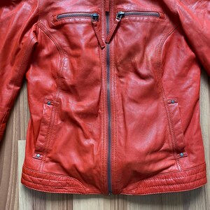 May include: A red leather jacket with a zip-up front, two zippered chest pockets, and two side pockets. The jacket has a classic biker style with a tailored fit and a stand-up collar.
