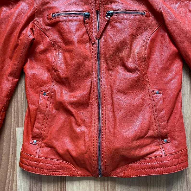 May include: A red leather jacket with a zip-up front, two zippered chest pockets, and two side pockets. The jacket has a classic biker style with a tailored fit and a stand-up collar.