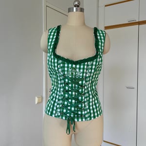 May include: A green and white checked corset top with a square neckline and a lace-up front. The top features green trim and a floral design near the neckline. The garment is displayed on a mannequin.
