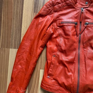 May include: A vibrant orange leather jacket with a quilted shoulder design and a zippered front. The jacket features two zippered pockets and a stand-up collar. The jacket is displayed on a wooden surface.