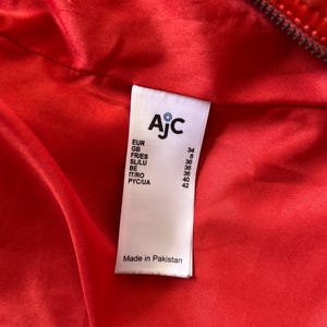 May include: Close-up of a vibrant red garment's interior, revealing a label with the brand name "AjC" and size conversions for various regions. The label also states "Made in Pakistan".