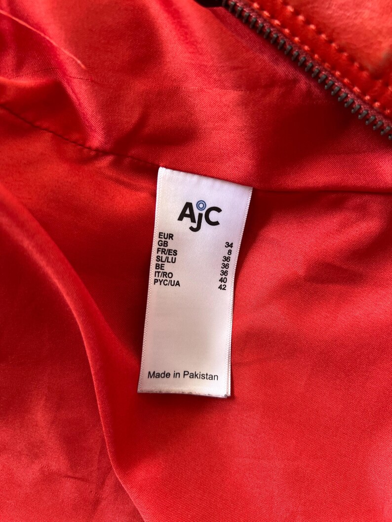 May include: Close-up of a vibrant red garment's interior, revealing a label with the brand name "AjC" and size conversions for various regions. The label also states "Made in Pakistan".