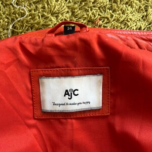 May include: Close-up of the inside of a red leather jacket. The lining is red, and a white label with the text "AjC Designed to make you happy" is sewn into the jacket. The size tag reads "34".