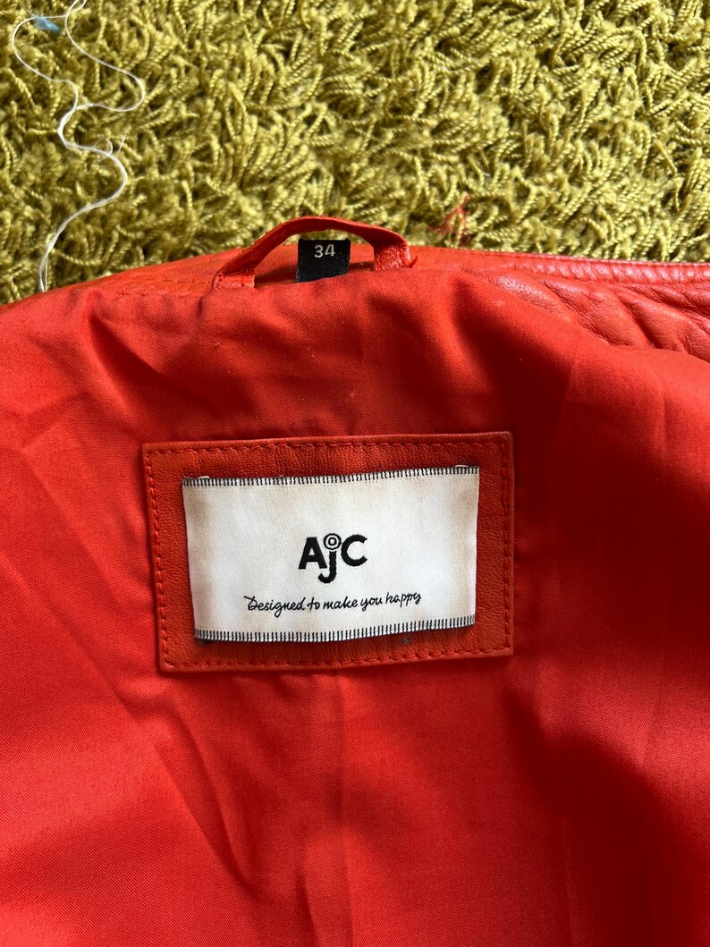 May include: Close-up of the inside of a red leather jacket. The lining is red, and a white label with the text "AjC Designed to make you happy" is sewn into the jacket. The size tag reads "34".