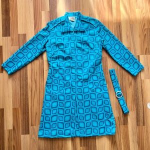 Op de afbeelding: Een fel turquoise jas met een herhalend geometrisch vierkantpatroon in donkerblauw, voorzien van een puntige kraag, lange mouwen, twee voorzakken en een ritssluiting over de gehele lengte. Een bijpassende stoffen riem met zilverkleurige gesp is inbegrepen.