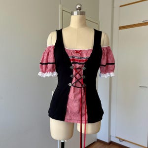 May include: Black and red gingham top with off-the-shoulder sleeves. The bodice has a corset-style front with red ribbon lacing and silver metal eyelets. The sleeves are red gingham with white lace trim.