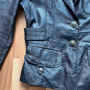K&ouml;nnte beinhalten: Nahaufnahme einer schwarzen Lederjacke mit dekorativen N&auml;hten und zwei Vordertaschen. Die Jacke hat zwei verzierte Kn&ouml;pfe und ein gekerbtes Revers. Das Leder scheint abgenutzt zu sein, was ihm eine Vintage-&Auml;sthetik verleiht.
