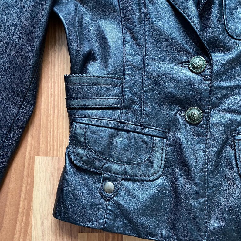 K&ouml;nnte beinhalten: Nahaufnahme einer schwarzen Lederjacke mit dekorativen N&auml;hten und zwei Vordertaschen. Die Jacke hat zwei verzierte Kn&ouml;pfe und ein gekerbtes Revers. Das Leder scheint abgenutzt zu sein, was ihm eine Vintage-&Auml;sthetik verleiht.
