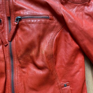 May include: Close-up of a red leather jacket. The jacket features a zippered front, a small zippered chest pocket, and a snap closure at the bottom. The leather has a slightly distressed appearance, with visible stitching and paneling details.