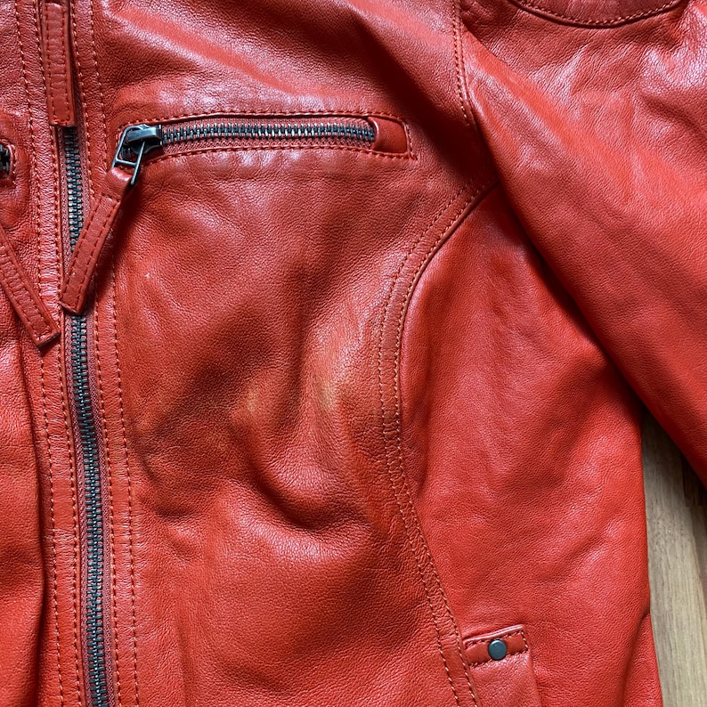May include: Close-up of a red leather jacket. The jacket features a zippered front, a small zippered chest pocket, and a snap closure at the bottom. The leather has a slightly distressed appearance, with visible stitching and paneling details.