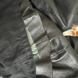 K&ouml;nnte beinhalten: Nahaufnahme einer schwarzen Lederjacke, die die Textur und die Nahtdetails zeigt. Die Jacke scheint aus hochwertigem Leder gefertigt zu sein, mit sichtbaren N&auml;hten und einem strukturierten Design. Das Bild hebt den Kragen und die &Auml;rmel hervor.