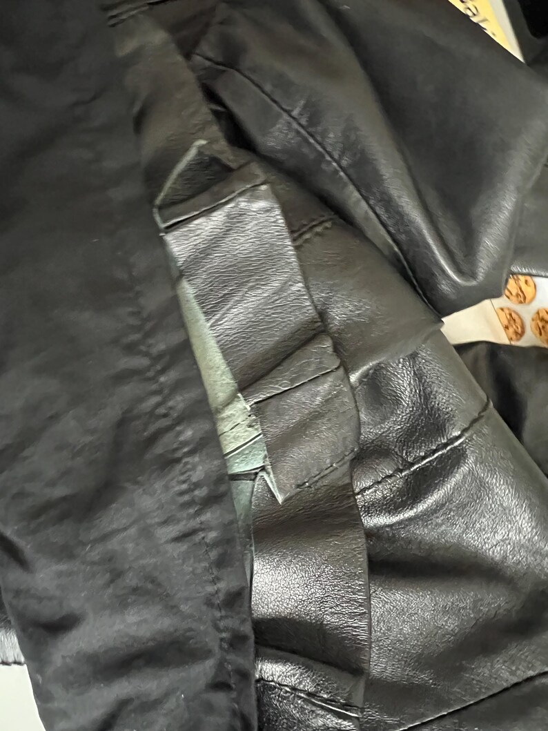 K&ouml;nnte beinhalten: Nahaufnahme einer schwarzen Lederjacke, die die Textur und die Nahtdetails zeigt. Die Jacke scheint aus hochwertigem Leder gefertigt zu sein, mit sichtbaren N&auml;hten und einem strukturierten Design. Das Bild hebt den Kragen und die &Auml;rmel hervor.