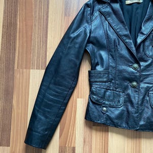 K&ouml;nnte beinhalten: Schwarze Lederjacke im Blazer-Stil. Sie hat einen Reverskragen, zwei Vordertaschen und Knopfverschl&uuml;sse. Die Jacke hat eine taillierte Form und einen leicht abgenutzten Look.