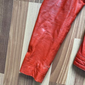 May include: Close-up of a red leather jacket sleeve. The sleeve is a vibrant shade of red, with visible stitching along the cuff and seams. The leather shows some wear and tear, adding character to the garment. The jacket appears to be vintage.