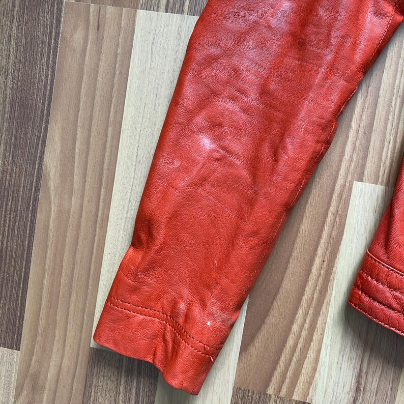 May include: Close-up of a red leather jacket sleeve. The sleeve is a vibrant shade of red, with visible stitching along the cuff and seams. The leather shows some wear and tear, adding character to the garment. The jacket appears to be vintage.