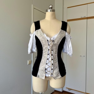 May include: A black and beige corset-style top with off-the-shoulder sleeves. The bodice has a lace-up front and decorative trim. The sleeves are white with lace edging. The top is displayed on a mannequin.