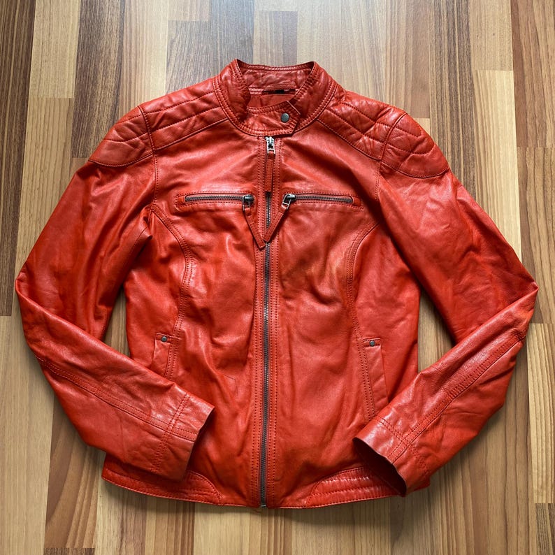 May include: A vibrant orange leather jacket with a quilted shoulder design and a stand-up collar. The jacket features a full-length zipper, two zippered chest pockets, and zippered cuffs. The jacket is a classic style.