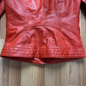 May include: A close-up of a red leather jacket. The jacket has a zipper closure and detailed stitching. The leather appears to be slightly textured. The jacket's design includes panels and seams, creating a tailored look.