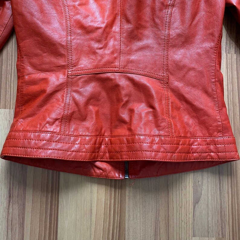 May include: A close-up of a red leather jacket. The jacket has a zipper closure and detailed stitching. The leather appears to be slightly textured. The jacket's design includes panels and seams, creating a tailored look.
