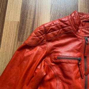May include: A close-up of a red leather jacket with a quilted shoulder design and a zippered pocket. The jacket has a stand-up collar and a front zipper closure. The leather has a textured appearance.