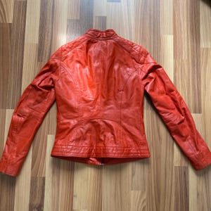 May include: A vibrant orange leather jacket with a stand-up collar and quilted shoulder detailing. The jacket has a zipper closure and seams that run down the back. The sleeves are long and the jacket is displayed on a wooden surface.