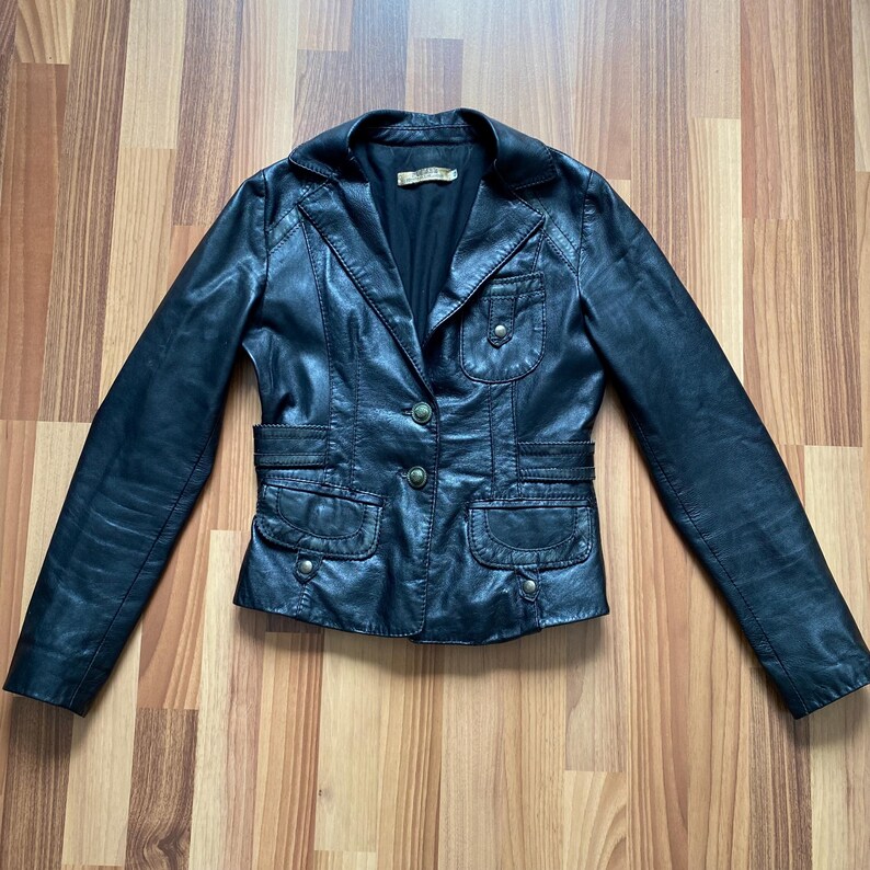 K&ouml;nnte beinhalten: Schwarze Lederjacke mit Reverskragen, Knopfverschluss und mehreren Taschen. Die Jacke hat eine taillierte Silhouette und ein klassisches Design. Die Jacke ist aus glattem, gl&auml;nzendem Leder gefertigt. Ein vielseitiges Kleidungsst&uuml;ck f&uuml;r jede Garderobe.