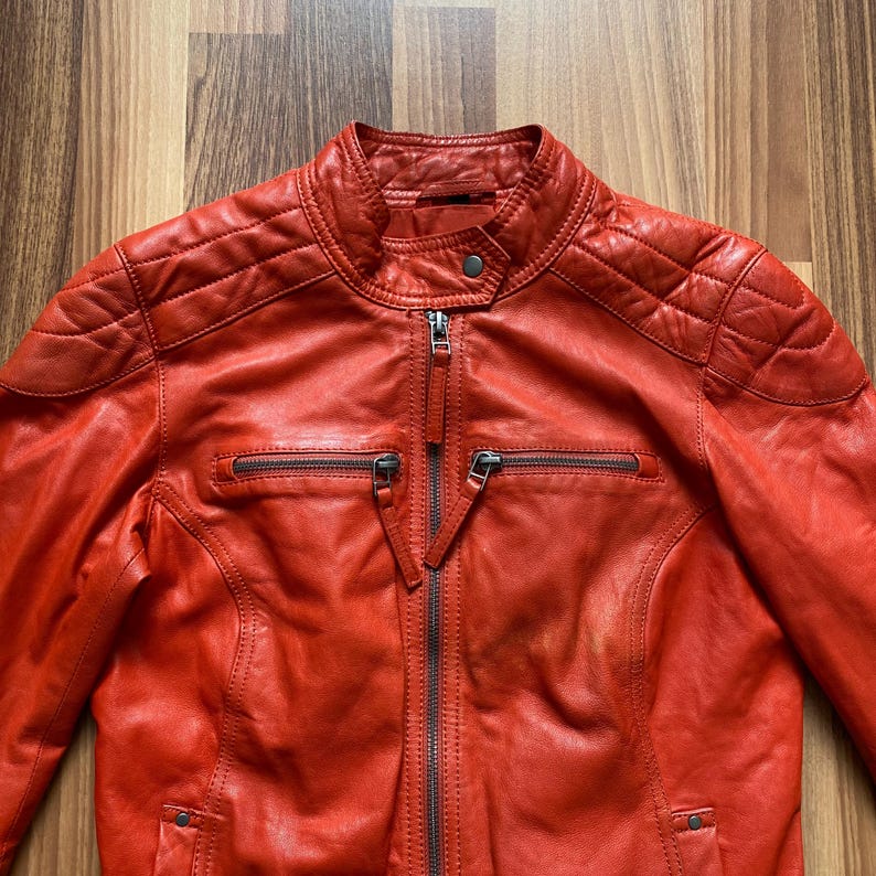 May include: A vibrant red leather jacket with a quilted shoulder design and a stand-up collar. The jacket features a full-length zipper, two zippered pockets, and a snap closure at the collar. The jacket is a classic style.
