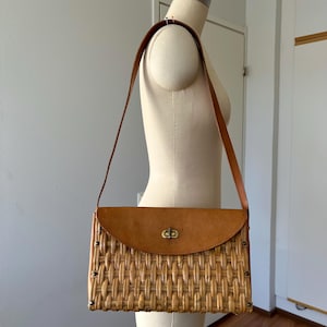 May include: A vintage rectangular handbag with a woven wicker base and a tan leather flap and strap. The flap is secured with a gold-coloured clasp. The bag is displayed on a mannequin.