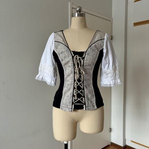 May include: A black and white corset-style top with a lace-up front. The bodice features a floral pattern and is trimmed with white lace. The sleeves are white with lace cuffs. The top is displayed on a mannequin.