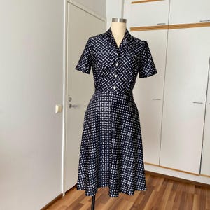 Könnte beinhalten: Ein knielanges Kleid aus dunkelblauem Stoff mit einem sich wiederholenden weißen geometrischen Muster. Das Kleid hat kurze Ärmel mit weißem Saum, einen Kragen, eine durchgehende Knopfleiste mit weißen Knöpfen und einen ausgestellten Rock.