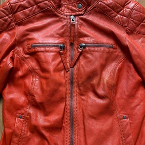 May include: A close-up of a red leather jacket with a zipper closure. The jacket has a quilted design on the shoulders and two zippered pockets on the chest. The leather has a slightly distressed appearance.