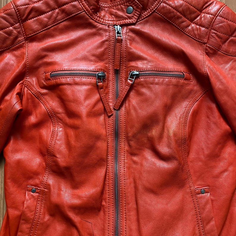 May include: A close-up of a red leather jacket with a zipper closure. The jacket has a quilted design on the shoulders and two zippered pockets on the chest. The leather has a slightly distressed appearance.