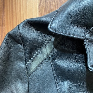 K&ouml;nnte beinhalten: Nahaufnahme von Kragen und Schulter einer schwarzen Lederjacke. Das Leder hat ein strukturiertes Aussehen mit sichtbaren N&auml;hten und einem kleinen Abnutzungsbereich. Die Jacke scheint Vintage oder abgenutzt zu sein.