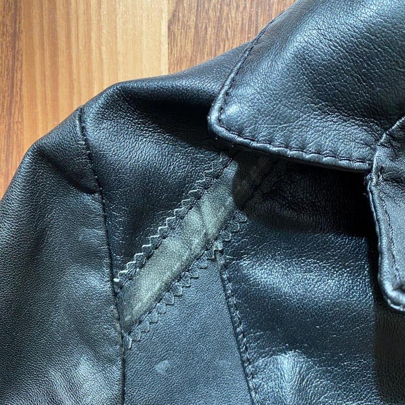 K&ouml;nnte beinhalten: Nahaufnahme von Kragen und Schulter einer schwarzen Lederjacke. Das Leder hat ein strukturiertes Aussehen mit sichtbaren N&auml;hten und einem kleinen Abnutzungsbereich. Die Jacke scheint Vintage oder abgenutzt zu sein.
