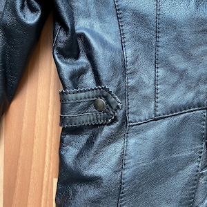 K&ouml;nnte beinhalten: Nahaufnahme einer schwarzen Lederjacke. Die Jacke hat detaillierte N&auml;hte, eine dekorative Klappe mit einem Knopf und eine strukturierte Oberfl&auml;che. Das Design deutet auf einen klassischen oder Vintage-Stil hin.